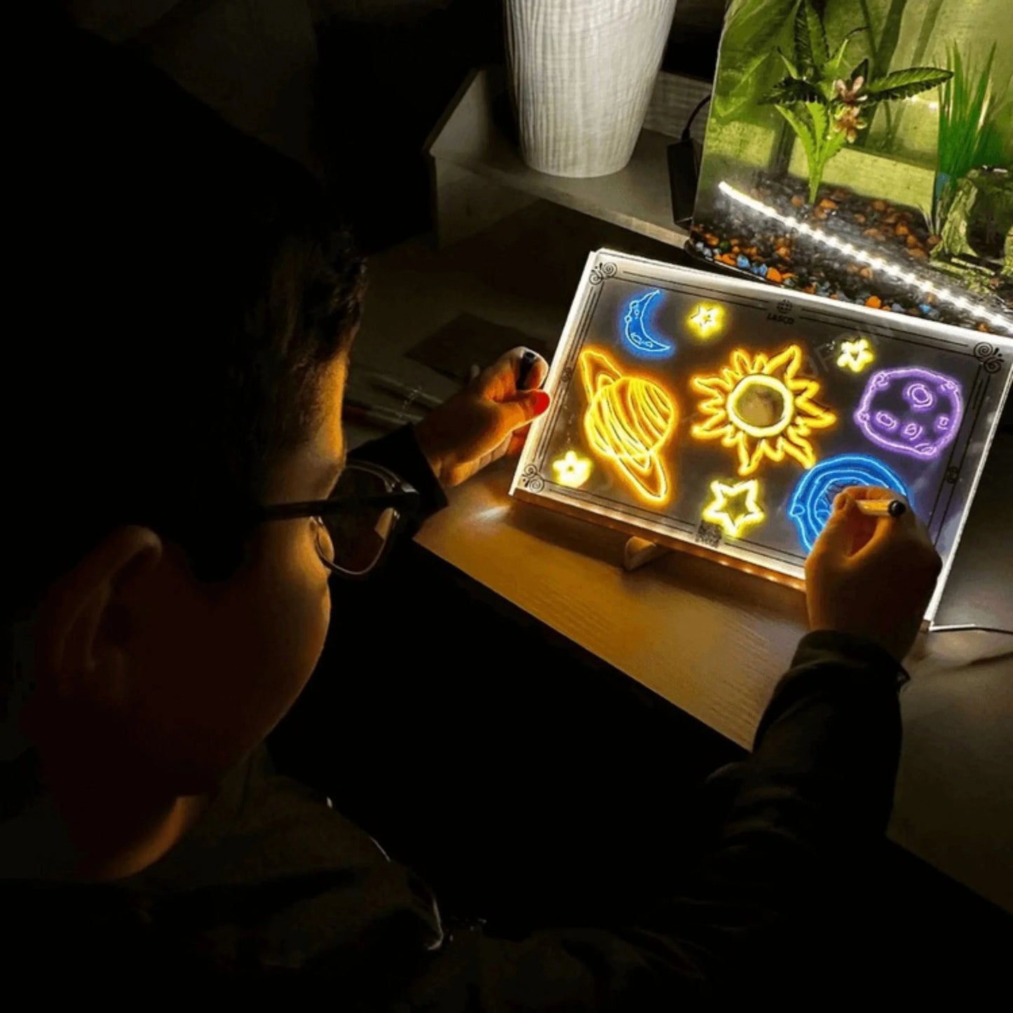 Criança brincando com lousa infantil iluminada, desenhando planetas e estrelas coloridos.